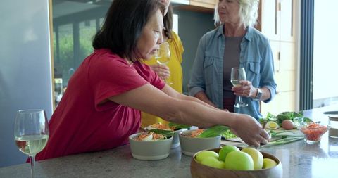 Diverse mature women preparing salads, sharing wine and conversation in sunlit kitchen