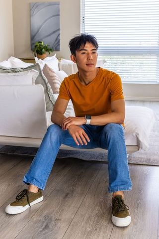 Young asian man sitting on bed edge wearing orange t-shirt in minimalist bedroom natural light