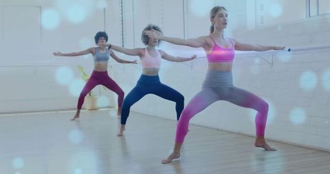 Instructor leading barre plié with trio of women practicing ballet fitness in sunlit studio