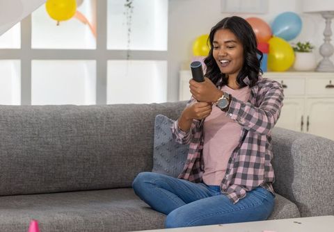 Indian woman celebrating at home twisting party popper on sofa with balloons and confetti