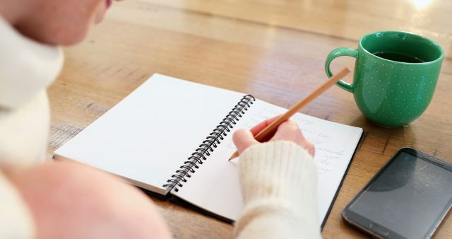 Young Caucasian woman writes in a notebook at home, with copy space. Cozy setting with a warm sweater, hot drink, and smartphone suggests planning or reflection.
