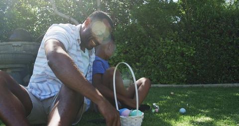 African american father and son arranging pastel easter basket on sunny backyard lawn