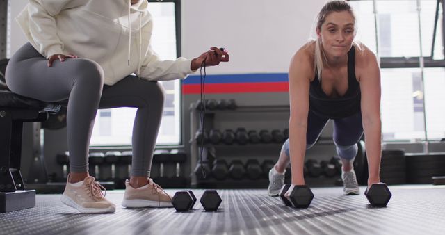 Image of diverse female fitness trainer timing determined woman raising dumbbells working out at gym. Exercise, fitness and healthy lifestyle.