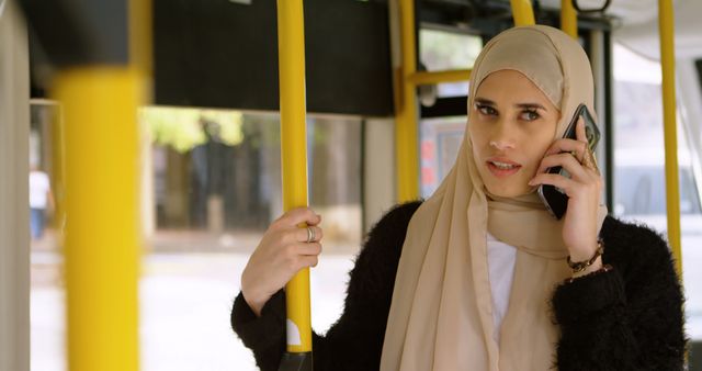 Muslim woman talks on the phone on a bus, with copy space. She's commuting, engaging in a conversation while traveling.