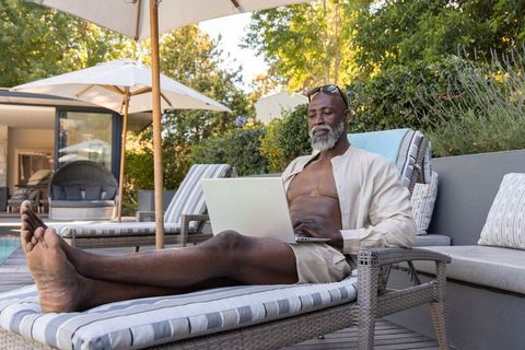 Senior african man reclining by pool using laptop, sunlit outdoor working and relaxing