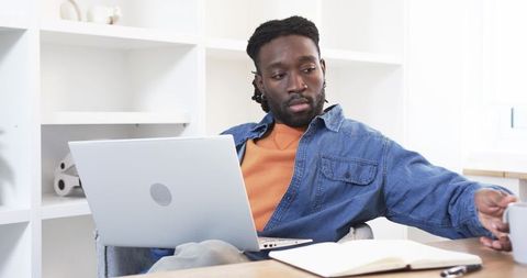 African American man working from home with laptop reaching for mug in bright home office