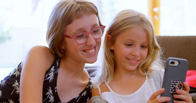 Teenage Caucasian girl and Caucasian girl enjoy a smartphone at home. They share a moment of joy as they engage with digital content, creating a warm, familial atmosphere.