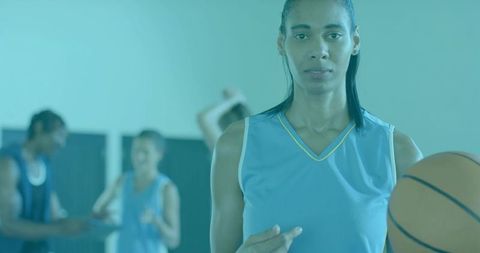 Female basketball player holding ball pointing at chest in gym during teal-lit team drill