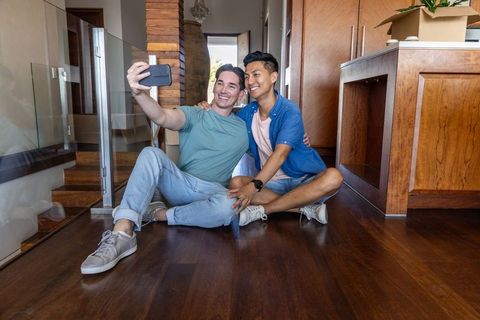 Caucasian and asian male partners moving in, taking selfie on hardwood floor near kitchen island