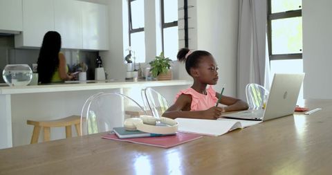 African American girl studying on laptop in modern kitchen while mother preparing breakfast