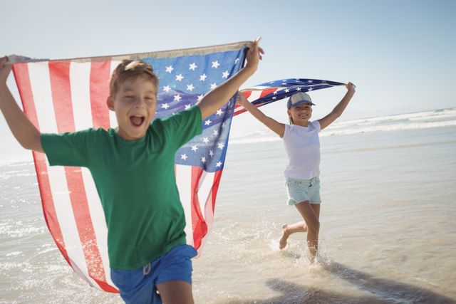 Happy siblings holding American flags while running on shore at beach during sunny day