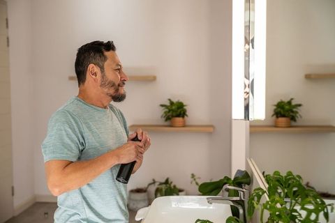 Mature asian man grooming at modern bathroom vanity holding black bottle looking at mirror
