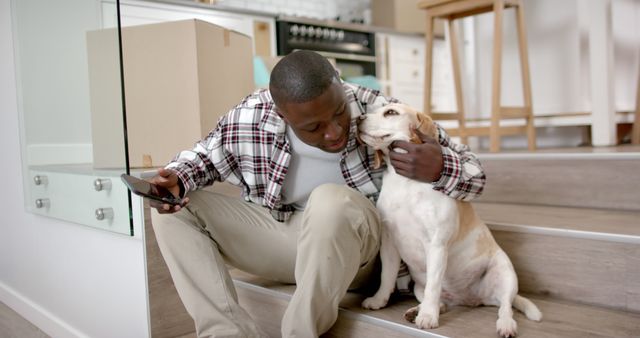 Man sitting on stairs in new home holding smartphone and cuddling dog. Ideal for themes related to relocation, pet ownership, lifestyle, modern apartments, companionship, and moving day. Perfect for blogs, articles, advertisements, and social media posts depicting new beginnings and pet-friendly living.