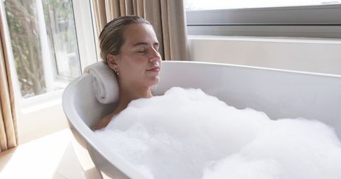 Woman relaxing in freestanding tub with bubble bath near sunlit window and rolled towel