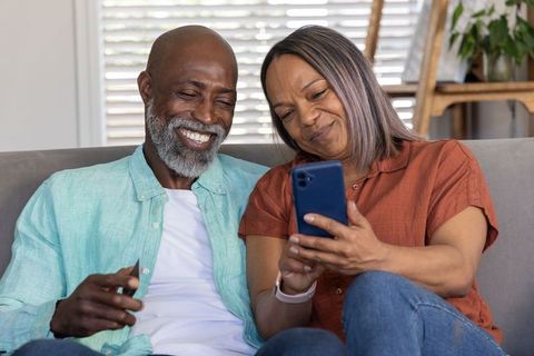 Senior couple sharing smartphone on living room couch, smiling and enjoying digital moment