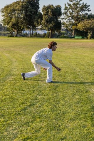 Crouching cricketer fielding on sunlit green outfield holding ball during golden hour