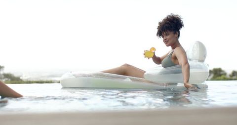 Relaxing woman lounging on inflatable raft in infinity pool holding tropical drink