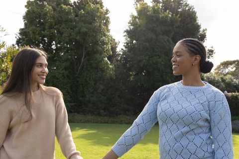 Smiling diverse women walking hand in hand on sunlit park lawn wearing knit sweaters
