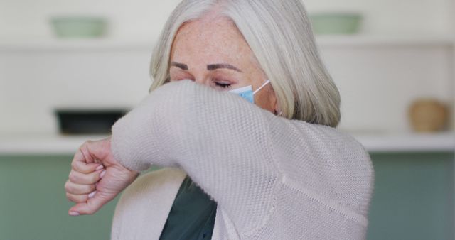 Elderly woman practicing good hygiene by coughing into her elbow while wearing a face mask. Useful for depicting health safety protocols, preventive actions, and promoting public health measures. Suitable for healthcare campaigns, educational materials, and articles on safety during illness.