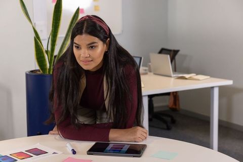 Asian woman leaning over round table reviewing tablet mockups and color palettes in office