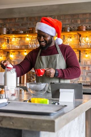 African american man wearing santa hat baking in rustic kitchen scooping sugar smiling