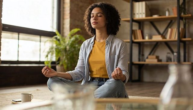 Young woman meditating cross-legged in loft living room wearing mustard top and jeans