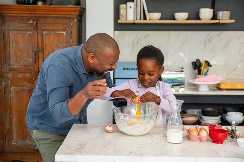 African american father laughing teaching daughter cracking egg mixing batter on marble countertop