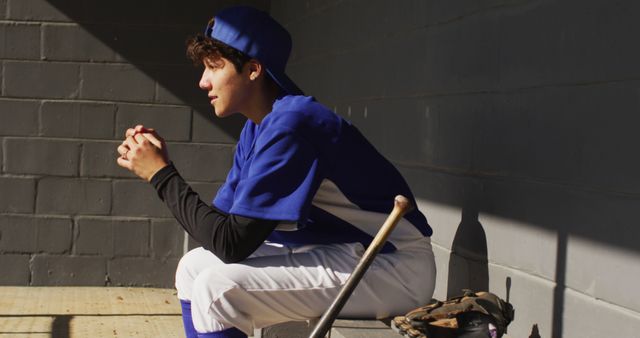 Nervous biracial female baseball player, sitting on bench in sun waiting with baseball bal. female baseball team, sports training and game tactics.