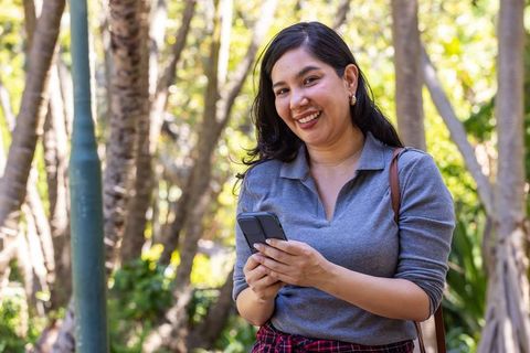Smiling woman holding smartphone in sunlit park wearing casual outfit and shoulder bag