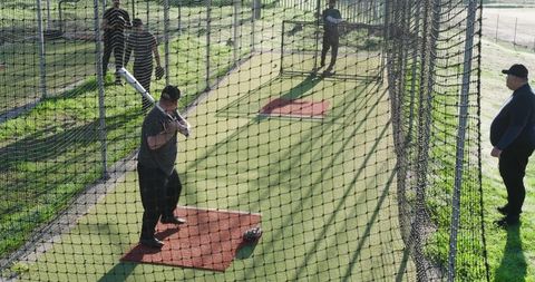 Teammates practicing batting in outdoor baseball cage under sunlit nets with coach