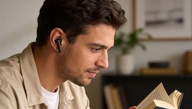 Young man reading with wireless earbuds in cozy living room, casual home study
