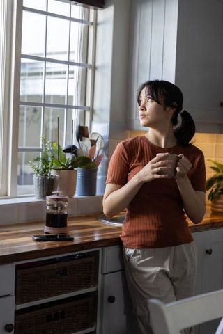 Asian woman holding mug by sunlit kitchen window, cozy morning routine, domestic calm