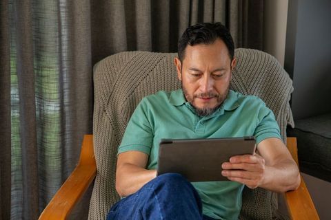 Mature asian man sitting in armchair holding tablet, enjoying home reading and browsing