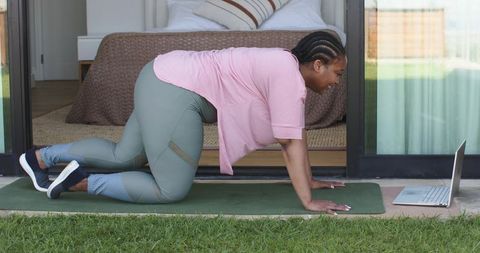 Mid-adult african american woman following online workout on patio mat with laptop