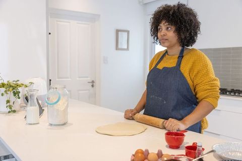 African american woman rolling dough on modern kitchen island wearing denim apron
