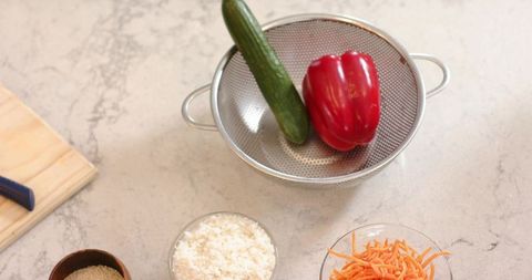 Stainless colander holding cucumber and red bell pepper with rice and shredded carrots