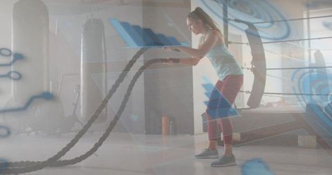 Female athlete powering battle ropes in boxing ring gym with double exposure technology overlay