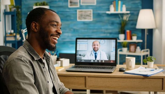 Smiling man consulting doctor via telehealth on laptop at home desk, showing medication
