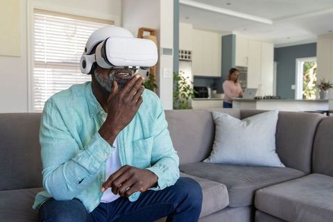 Black senior man wearing VR headset on sofa, partner working on laptop in modern home