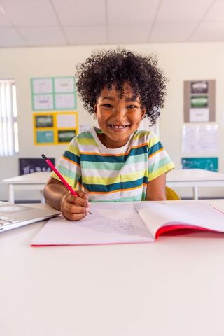 Smiling african american child writing in notebook holding red pencil at bright classroom desk