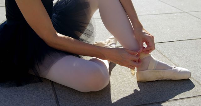 Ballerina ties her pointe shoes outdoors, with copy space. She prepares for a performance or practice in an urban setting.
