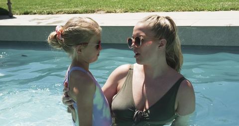 Mother and daughter bonding in backyard pool wearing sunglasses and summer swimsuits