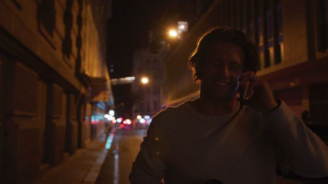 Night scene capturing a man enjoying the city atmosphere, chatting on his smartphone while holding a takeaway coffee. Ideal for concepts related to city lifestyle, nightlife, communication, and leisurely night strolls.