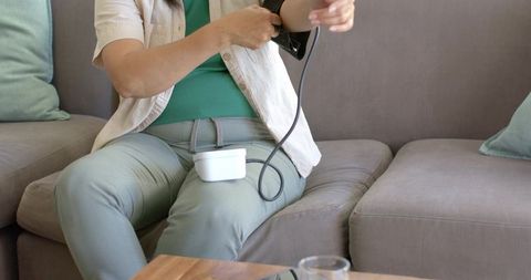 Senior woman checking blood pressure on gray sofa at home with portable monitor on lap