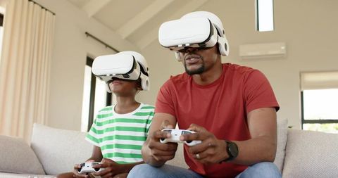 African American father and son playing virtual reality games on sofa, wearing VR headsets