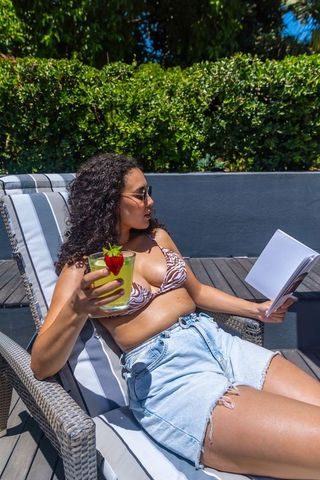 African american woman relaxing on lounge reading book and sipping strawberry cocktail
