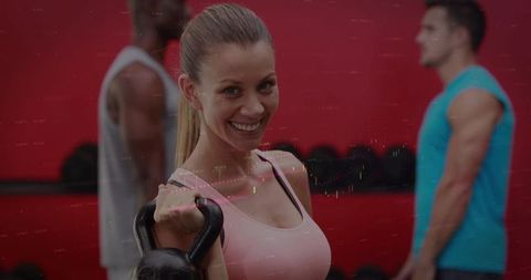 Smiling woman lifting kettlebell in gym wearing pink tank top during strength training