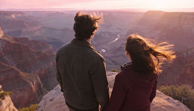 Couple standing on cliff gazing at sunset over layered canyon, windblown hair