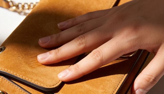 Resting feminine hand on tan suede clutch showing gold chain detail and leather texture