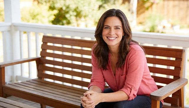 Smiling woman relaxing on porch bench, leaning forward clasping hands, casual coral shirt